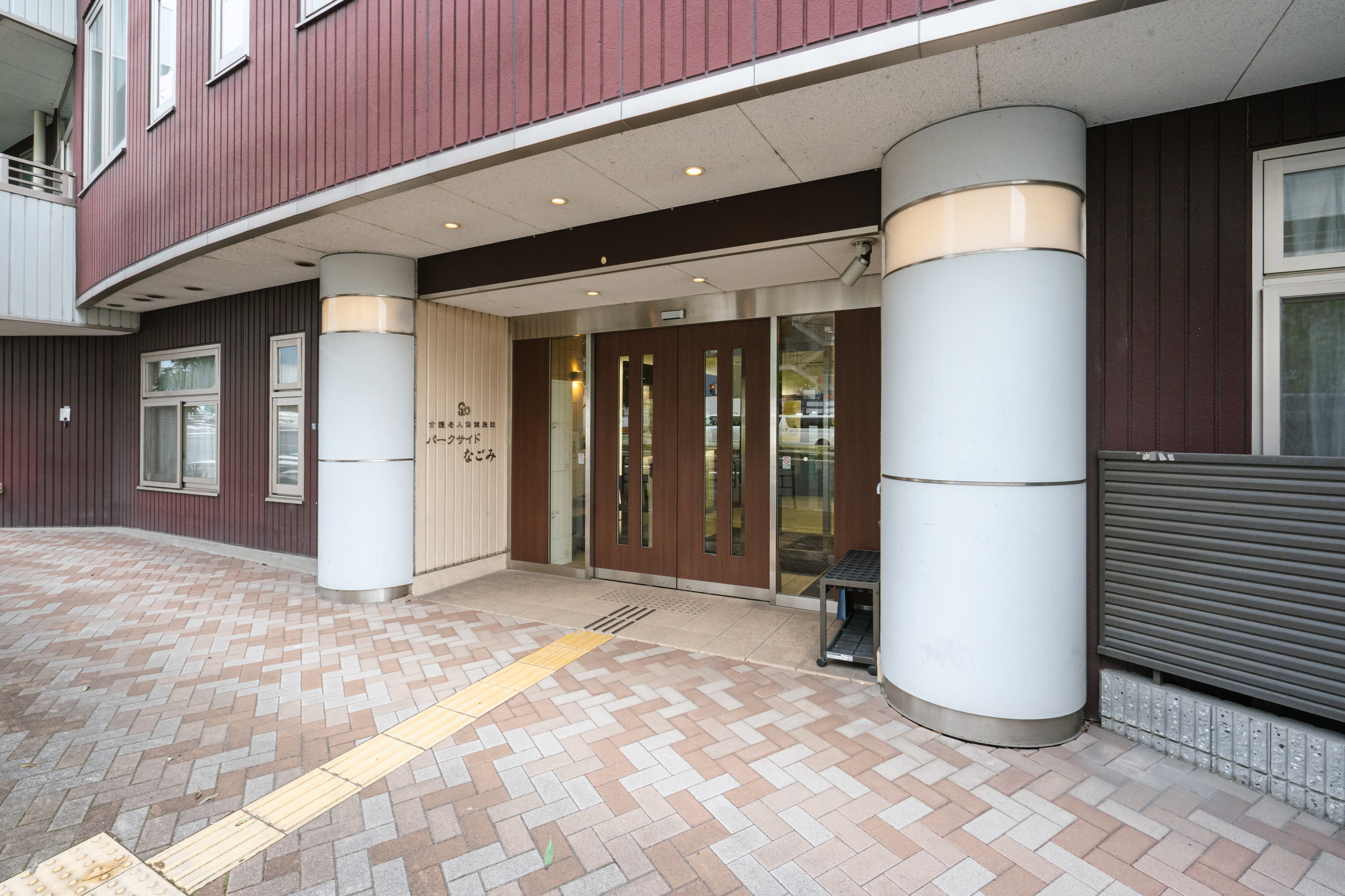 介護施設の玄関・車寄せ。赤茶色の外壁と白い円柱が特徴的で、レンガ舗装の前庭からアプローチしている。