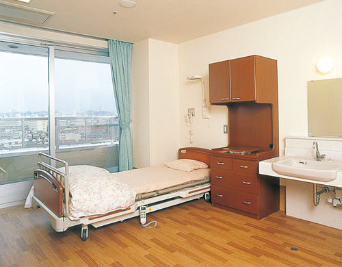 介護用ベッド、洗面台、収納家具を備えた居室。大きな窓から市街地が見える。